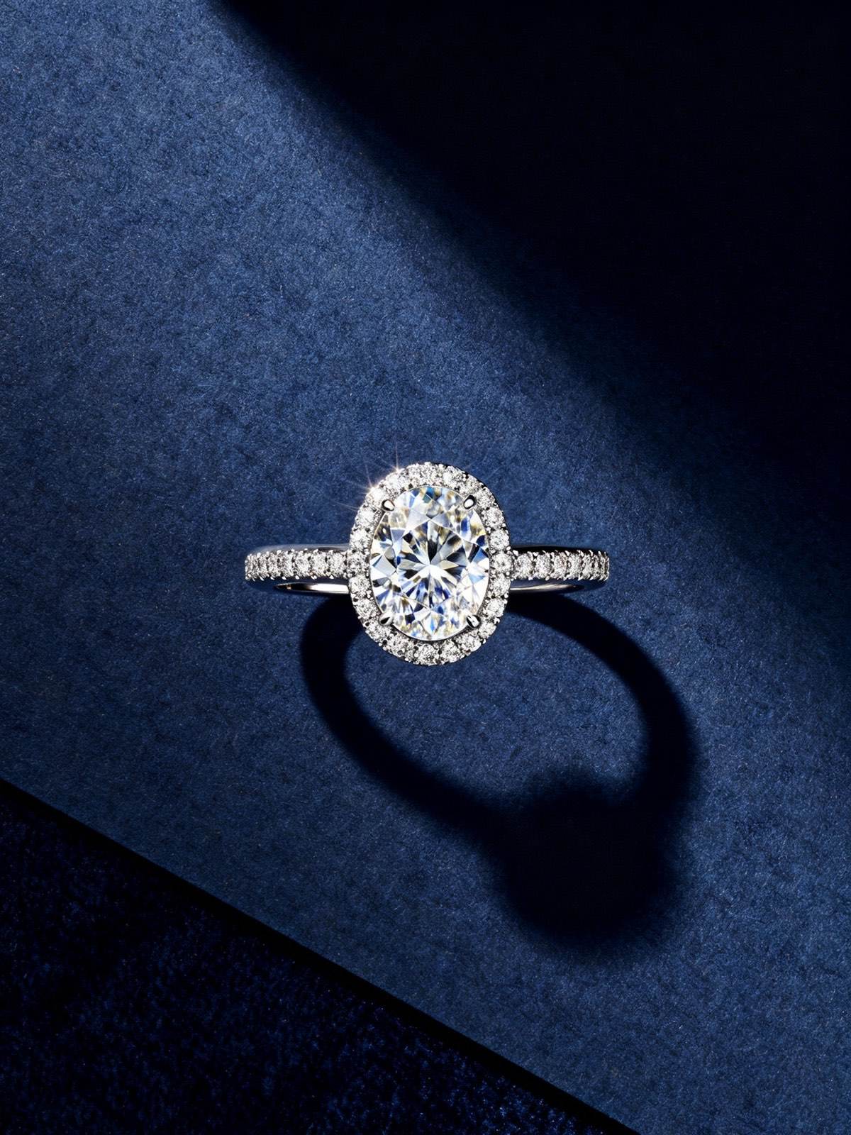 A woman's hand wearing a solitaire round brilliant engagement ring, editorial lighting on a midnight navy backdrop.